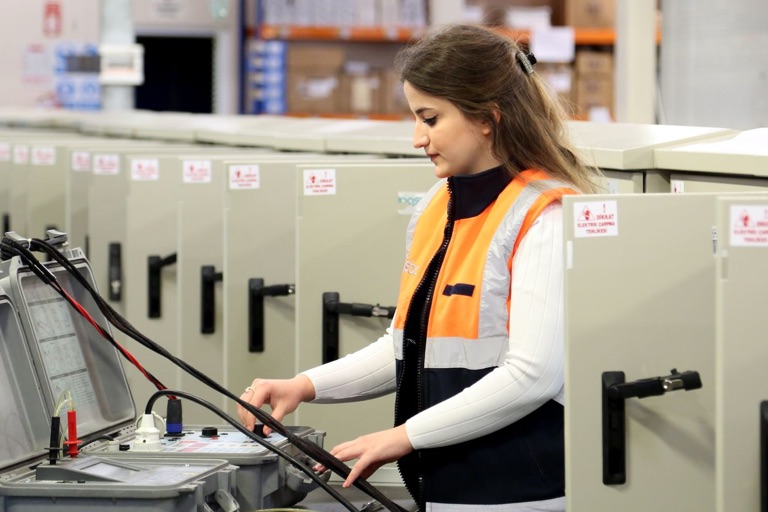 Woman electrical technician in a hi-vis vest testing control cabinets with an instrument